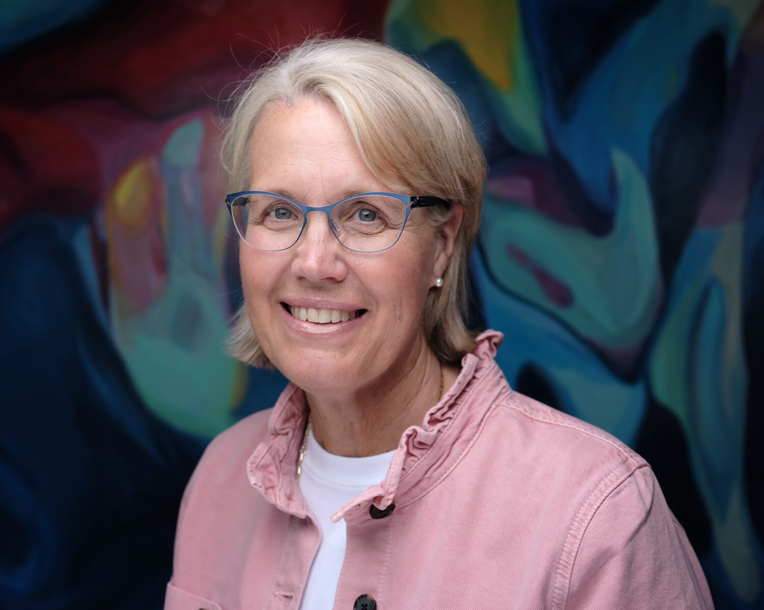 Pam Jensen wears a pink jacket and blue glasses. She has short blonde hair and smiles.