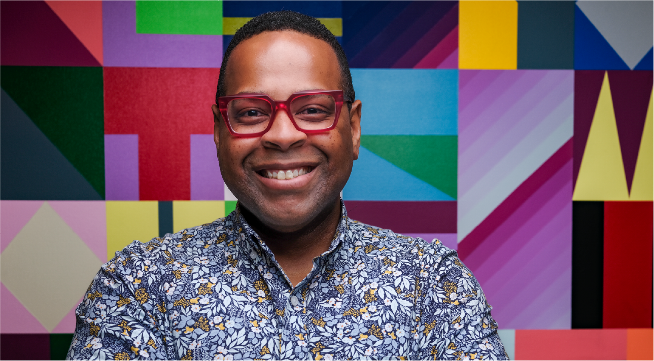 Adrian Jones wears red glasses and a floral collared shirt. He has close-cropped hair and smiles brightly.