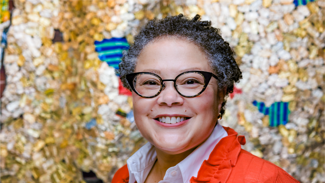 Kim Haskins wears a bright orange sweater and cat-eye glasses. She has short curly hair and smiles joyfully.