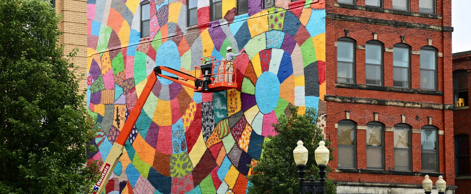 An artist in a cherry picker works on a large, colorful mural on the side of a building.