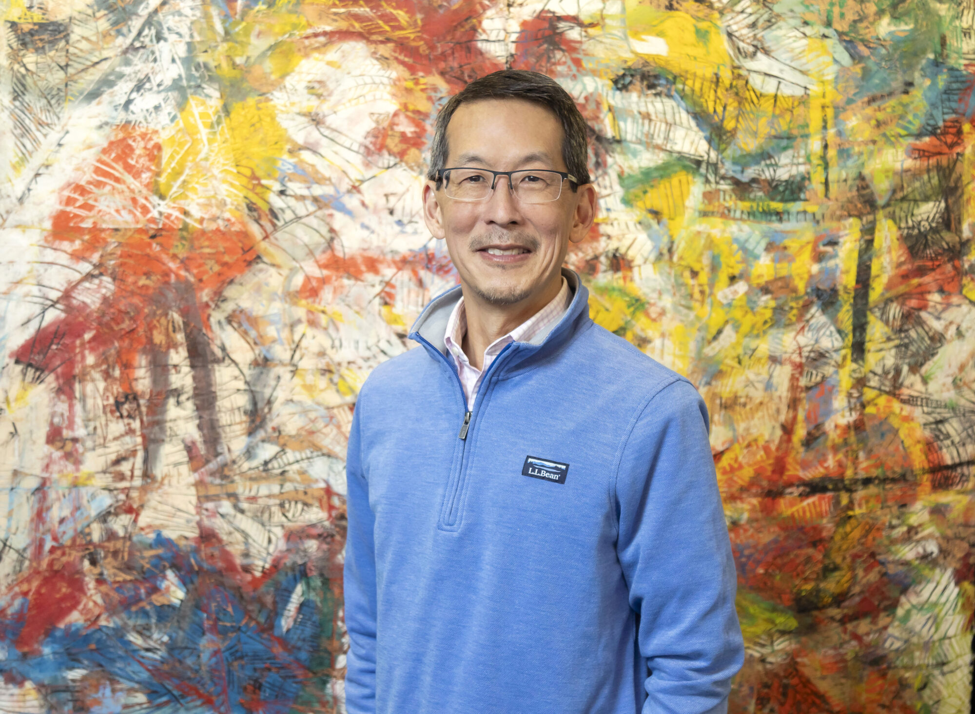 Roger Nozaki poses for a headshot. He wears a blue pullover and glasses and smiles warmly.