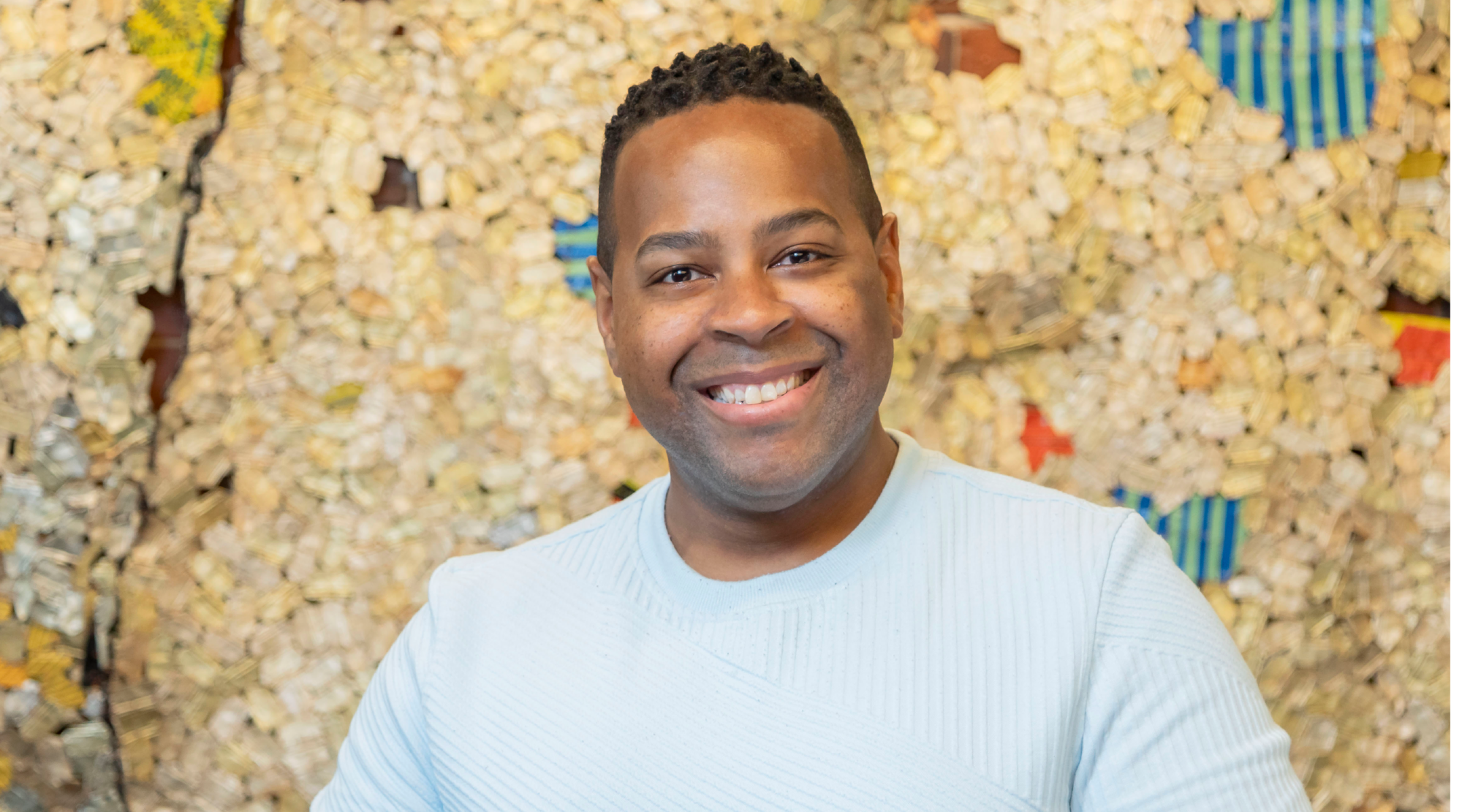Adrian Jones poses for a headshot. He wears a light blue sweater and smiles with joy.