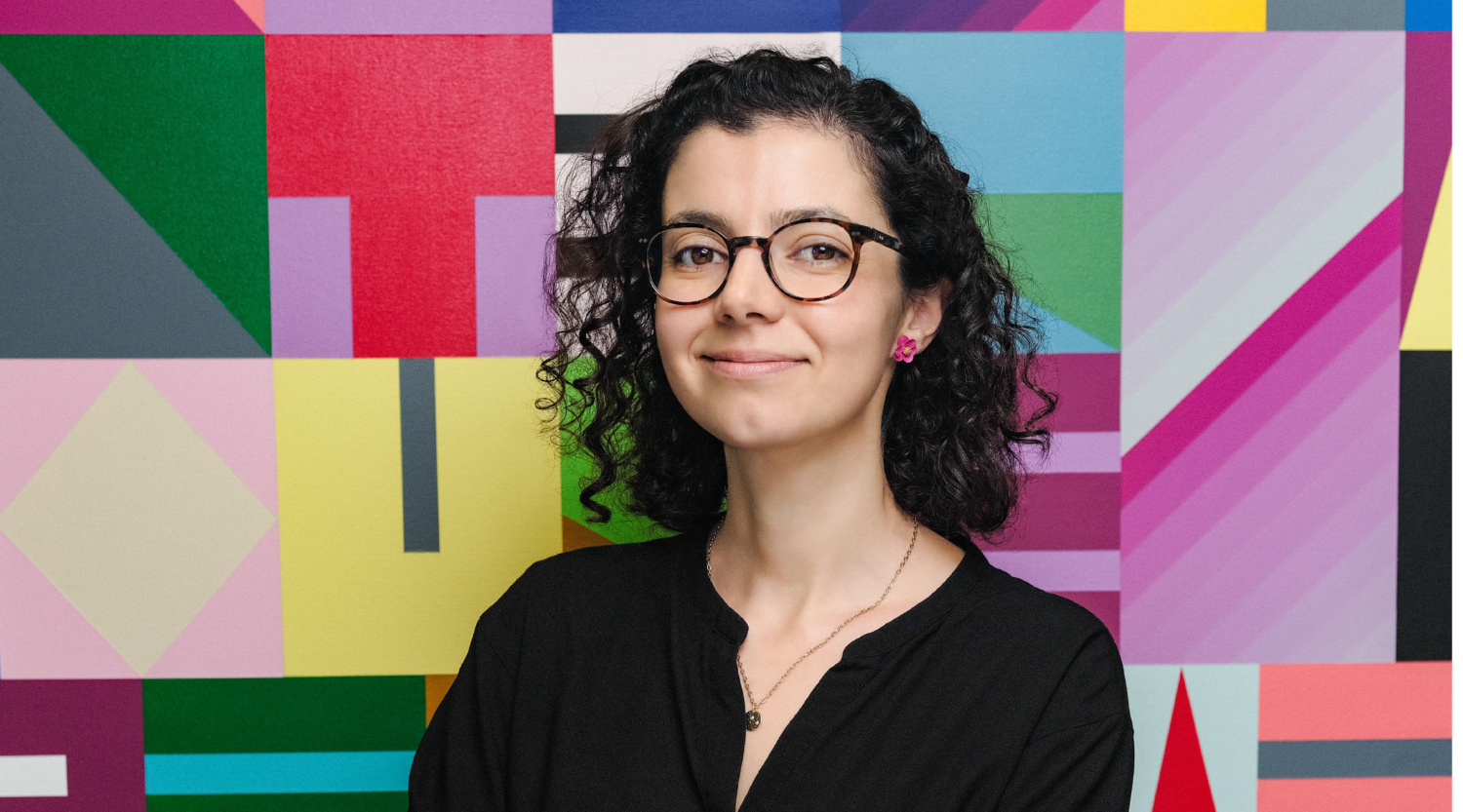 Andrea Prado Tuma poses for a headshot. She has black, shoulder length curly hair and wears glasses.