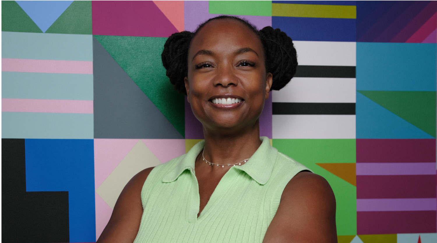 Erica Nunnally poses for a headshot. She wears s bright green top and styles her locs in two buns. She smiles with joy.
