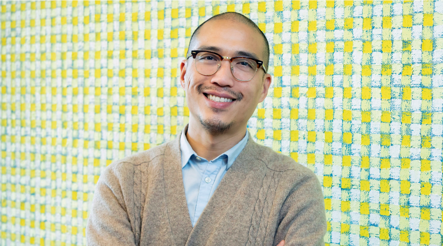 Giles Li poses for a headshot. He wears glasses and a neutral cardigan. He smiles warmly.