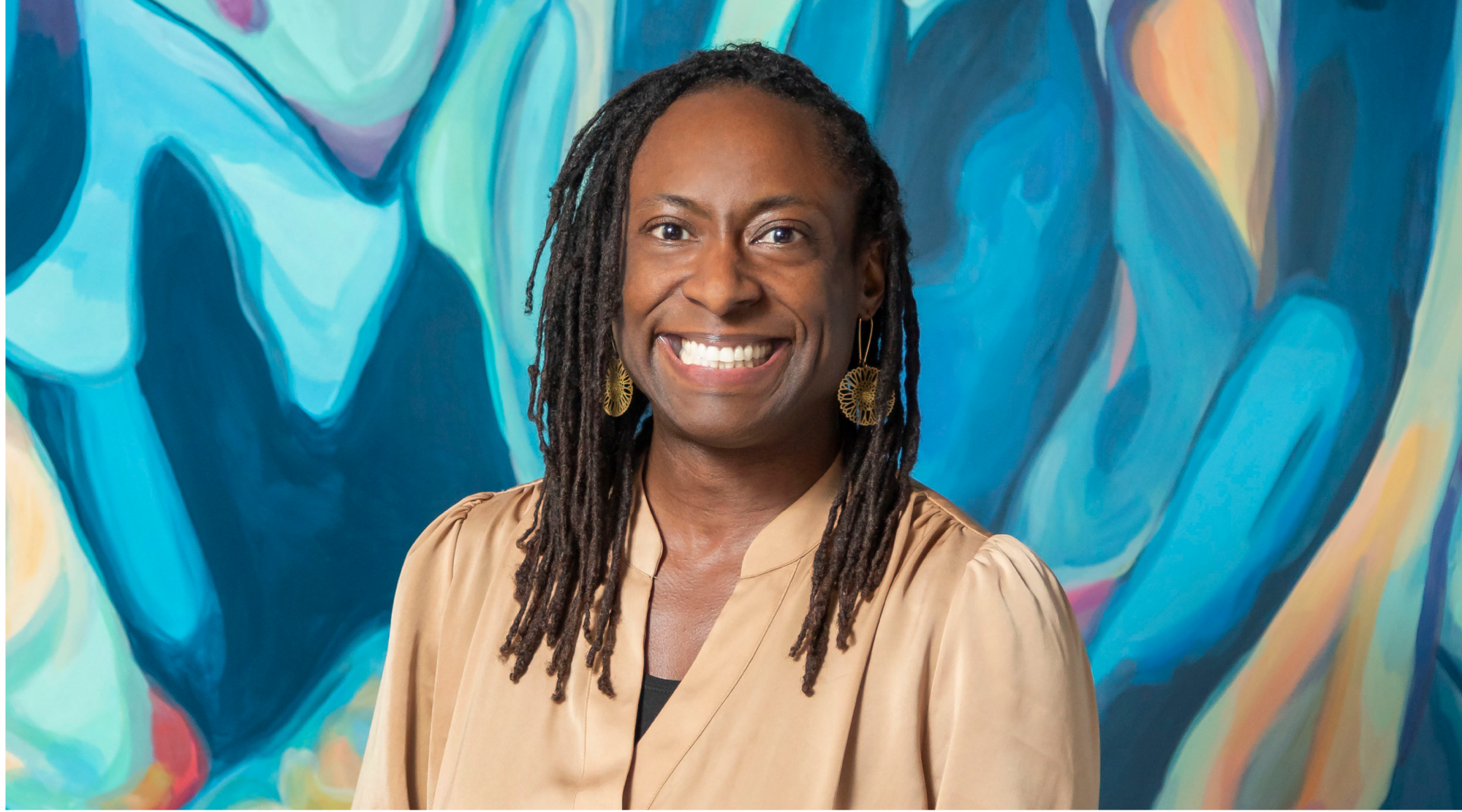 Jamilah Bradshaw poses for a headshot. She wears a silky gold top, and smiles joyfully.