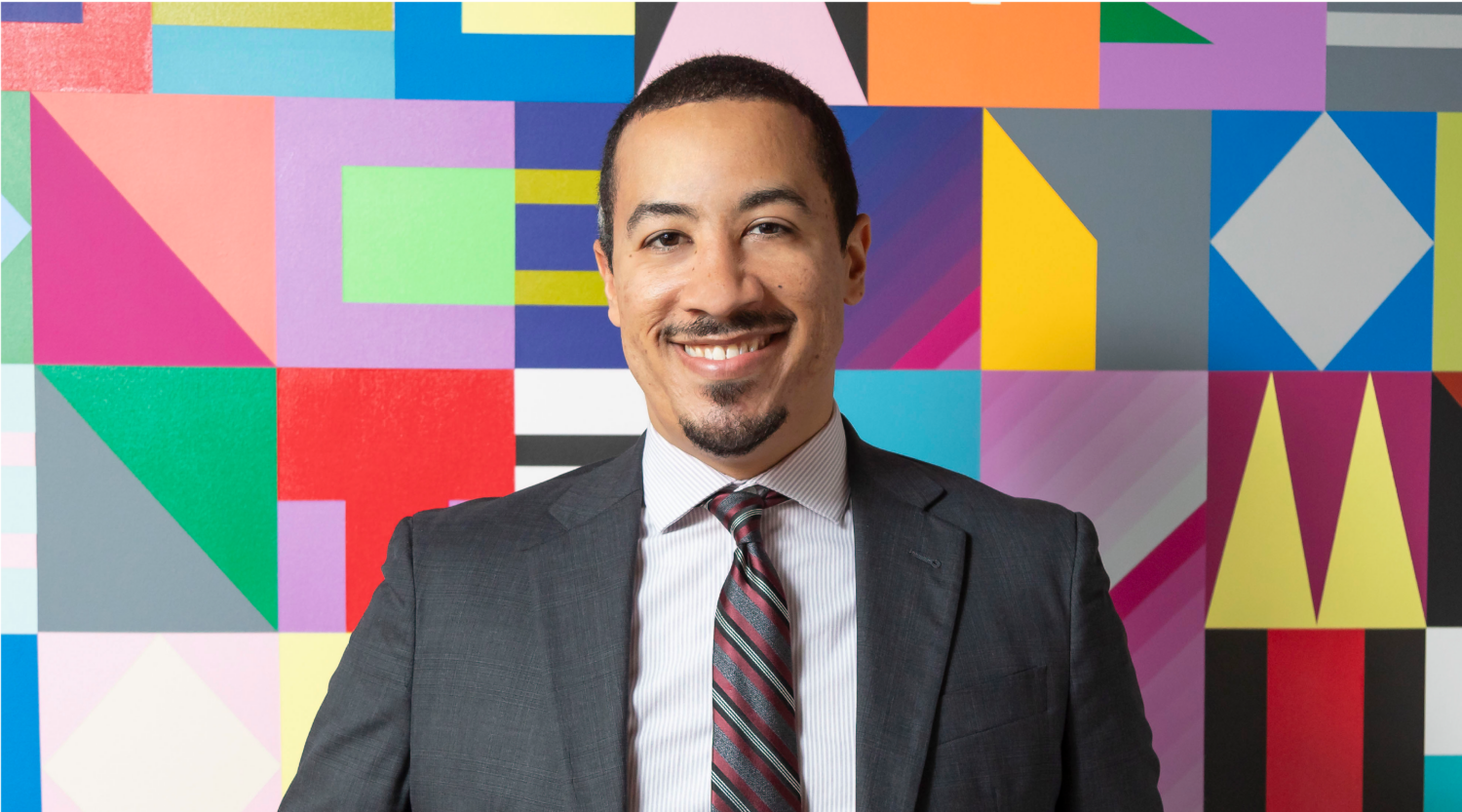 John Travis poses for a headshot. He wears a suit and smiles with joy.