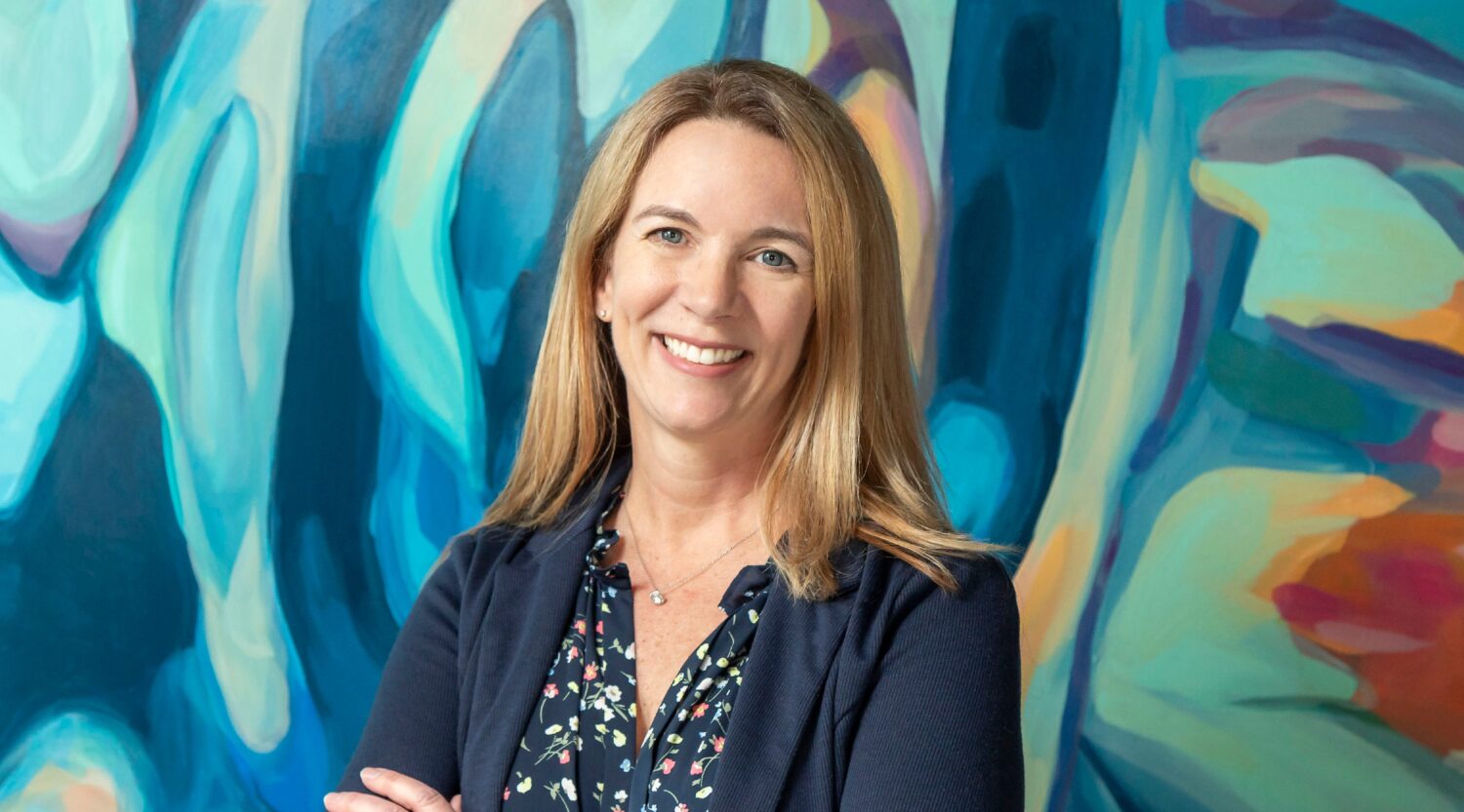 Kelly Perkins poses for a headshot. She has blonde hair and wears a denim jacket. She smiles warmly.