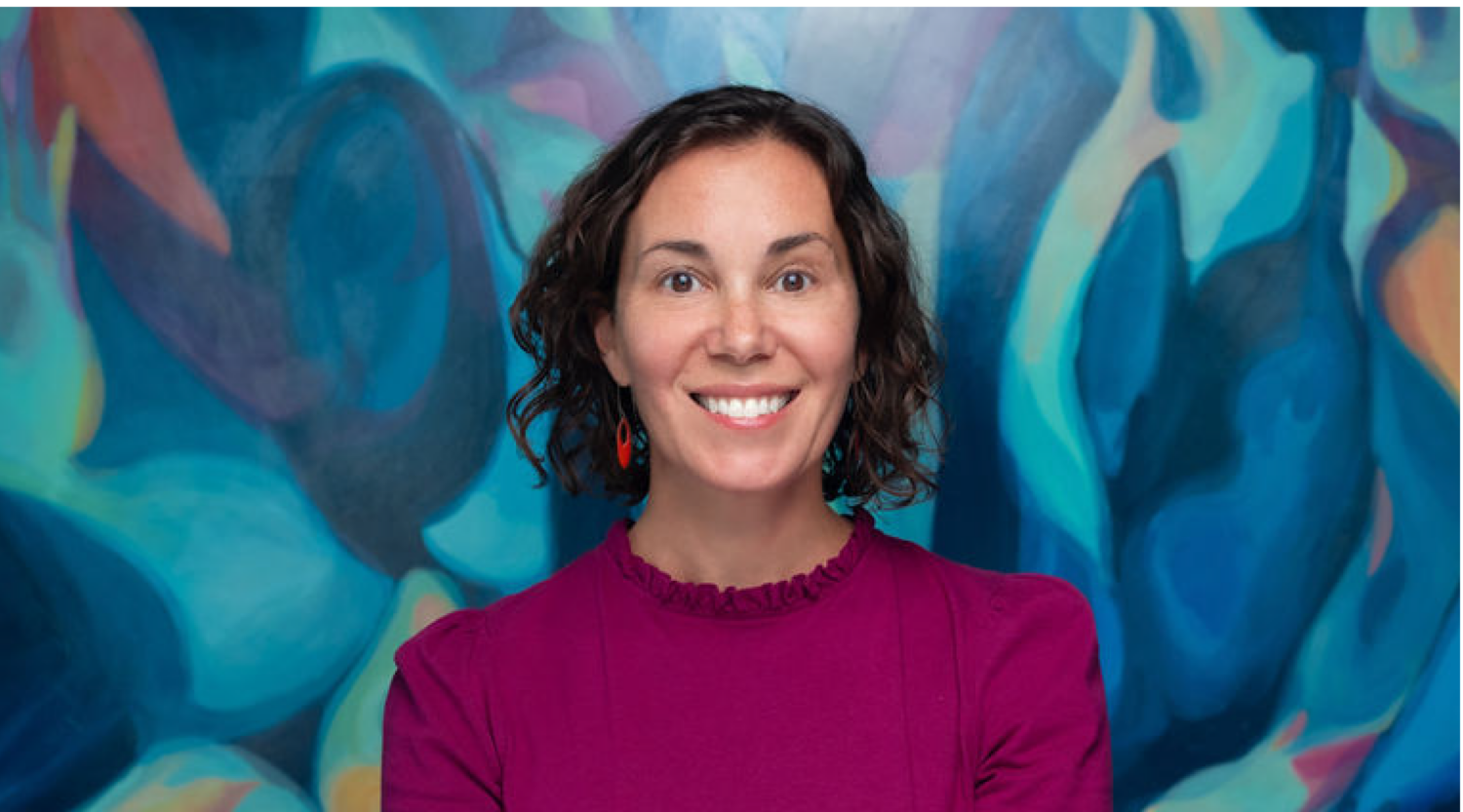 Lisa Jacobson poses for a headshot. She has shoulder-length wavy hair and wears a plum colored top. She smiles joyfully.