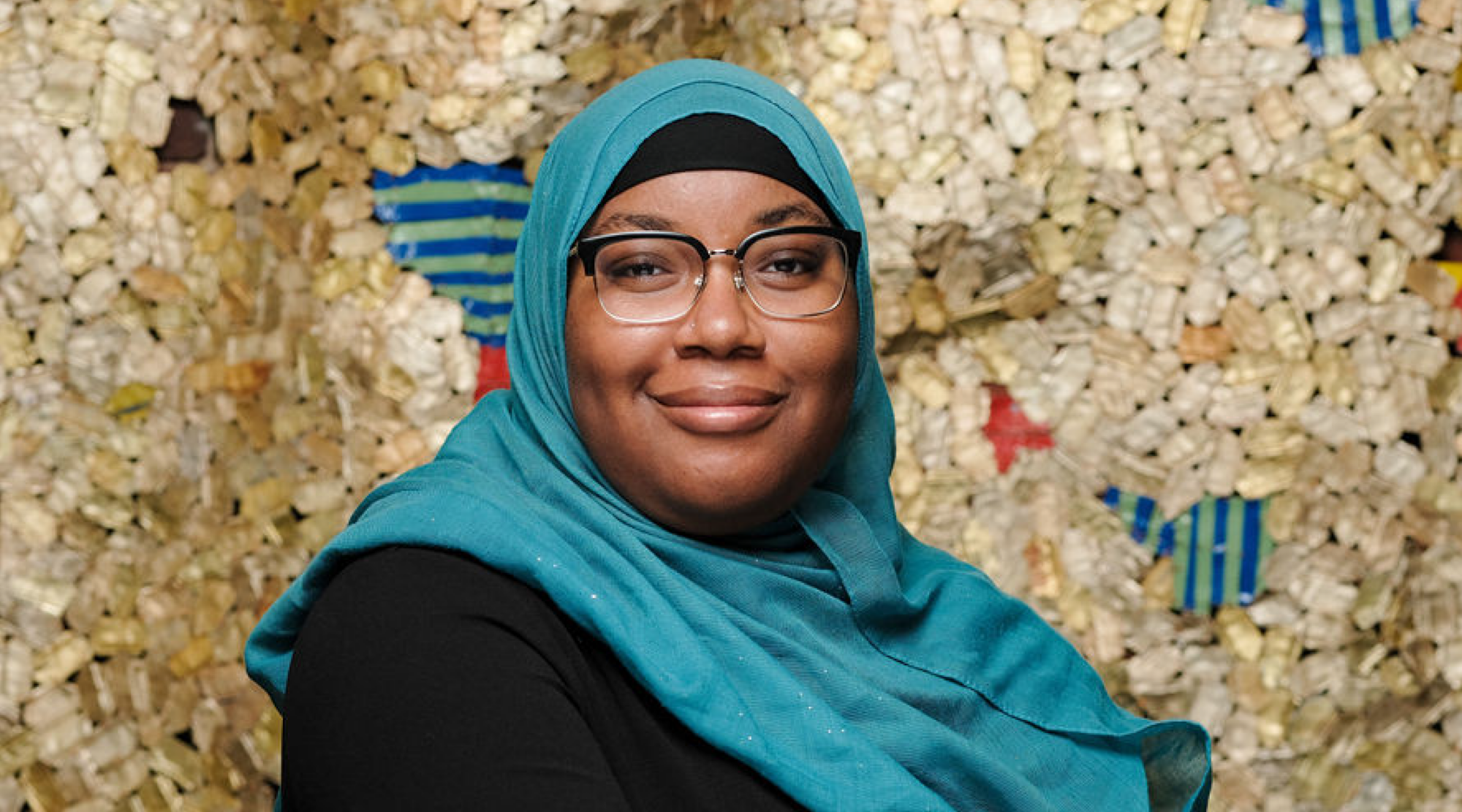 Najah Casimir poses for a headshot. They wear a bright teal hijab and a black dress. They smile warmly.