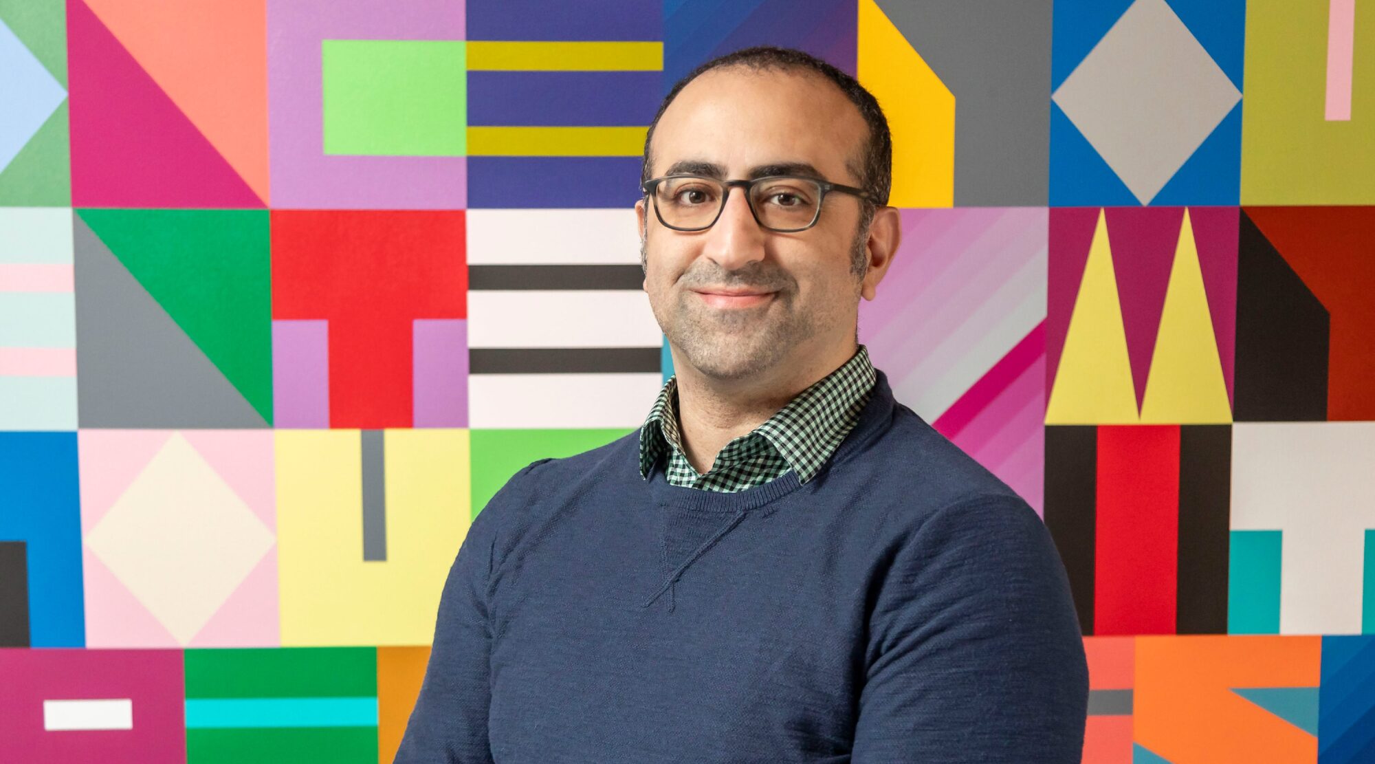 Pouya Shahbodagi poses for a headshot. He wears glasses, a collared shirt and sweater, and smiles warmly.