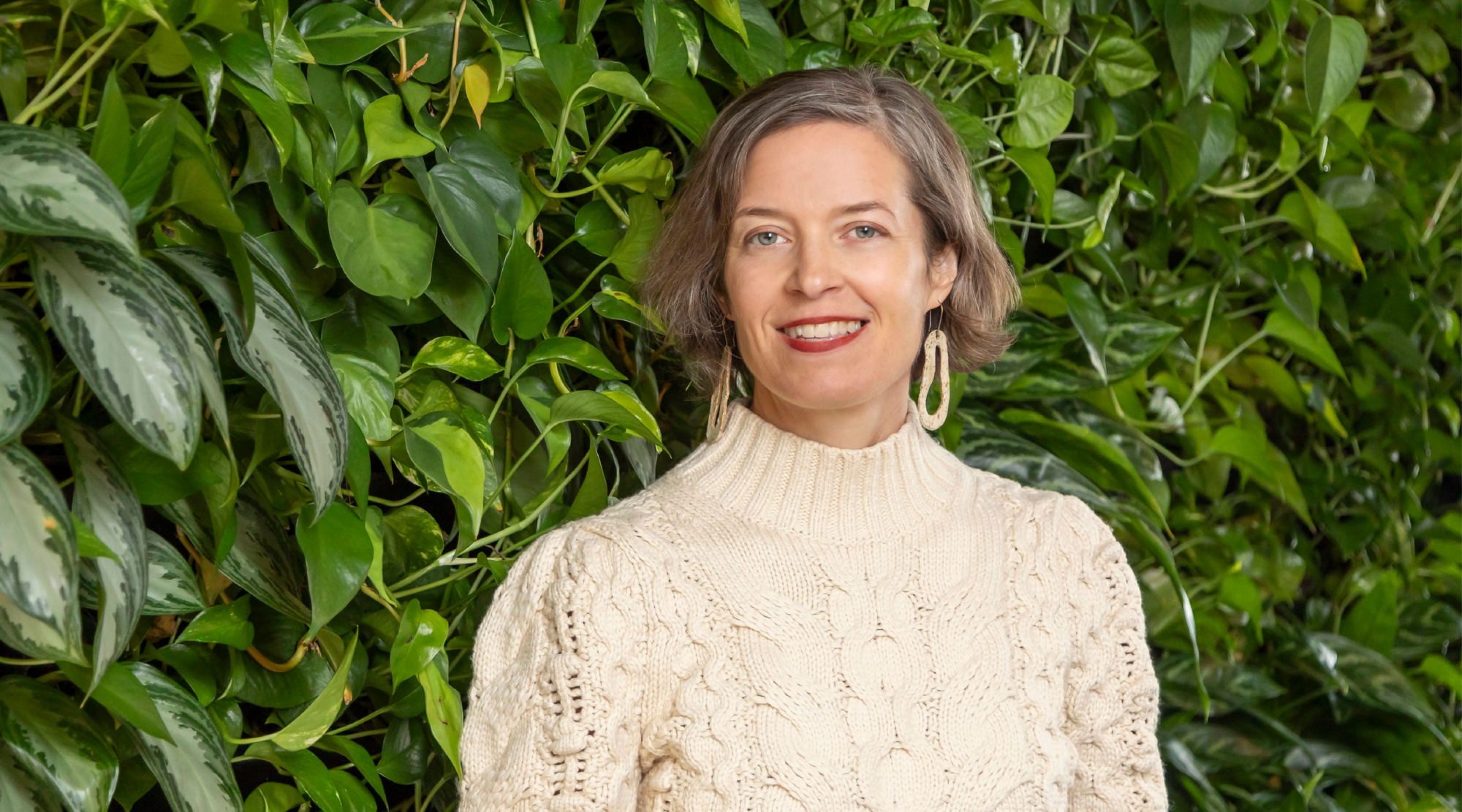 SueEllen Kroll poses for a headshot. She wears a white sweater and smiles in front of a wall of greenery.