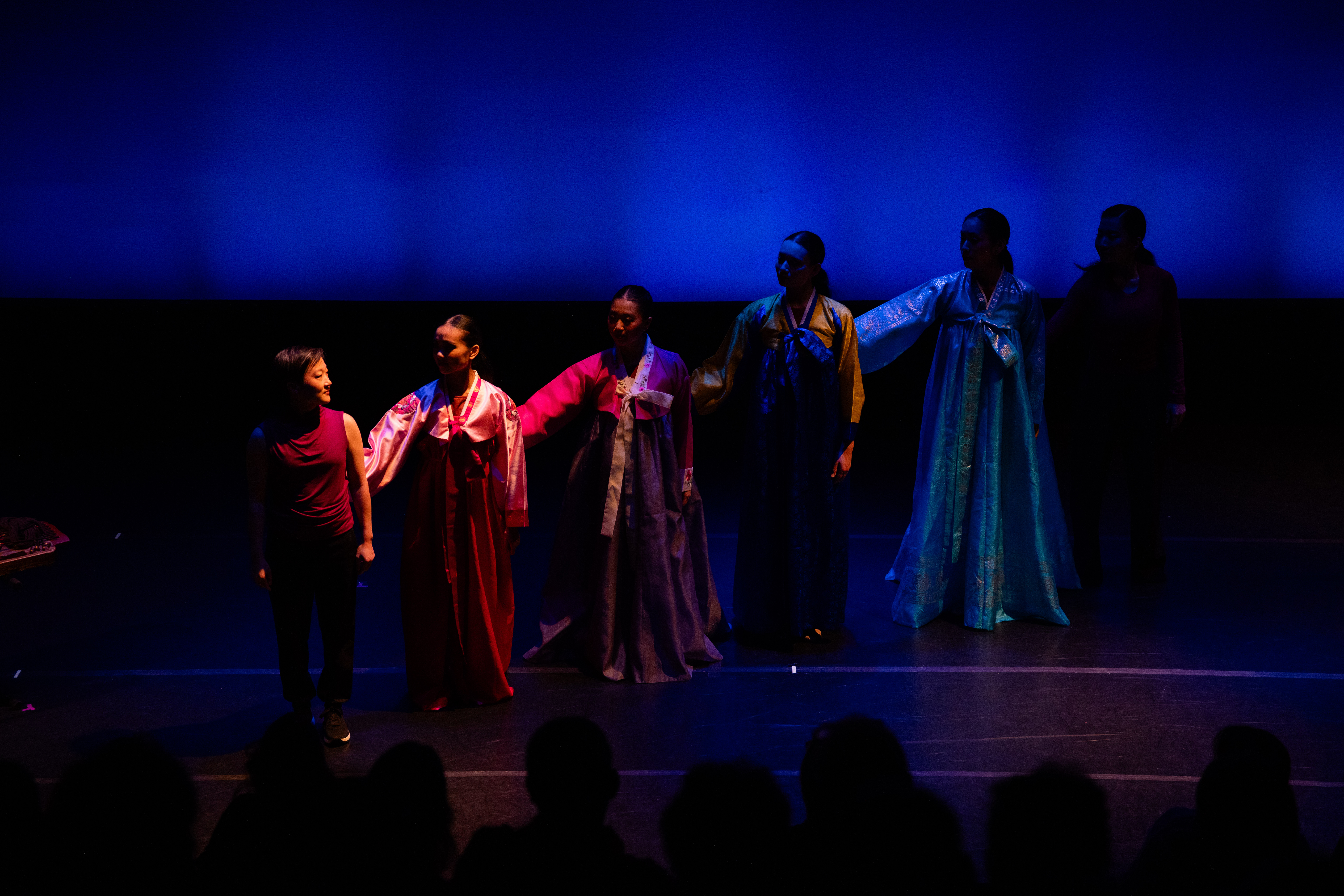 A group of dancers stand in a line across a dimly lit stage, hands on each others shoulders. They wear colorful silk robes.