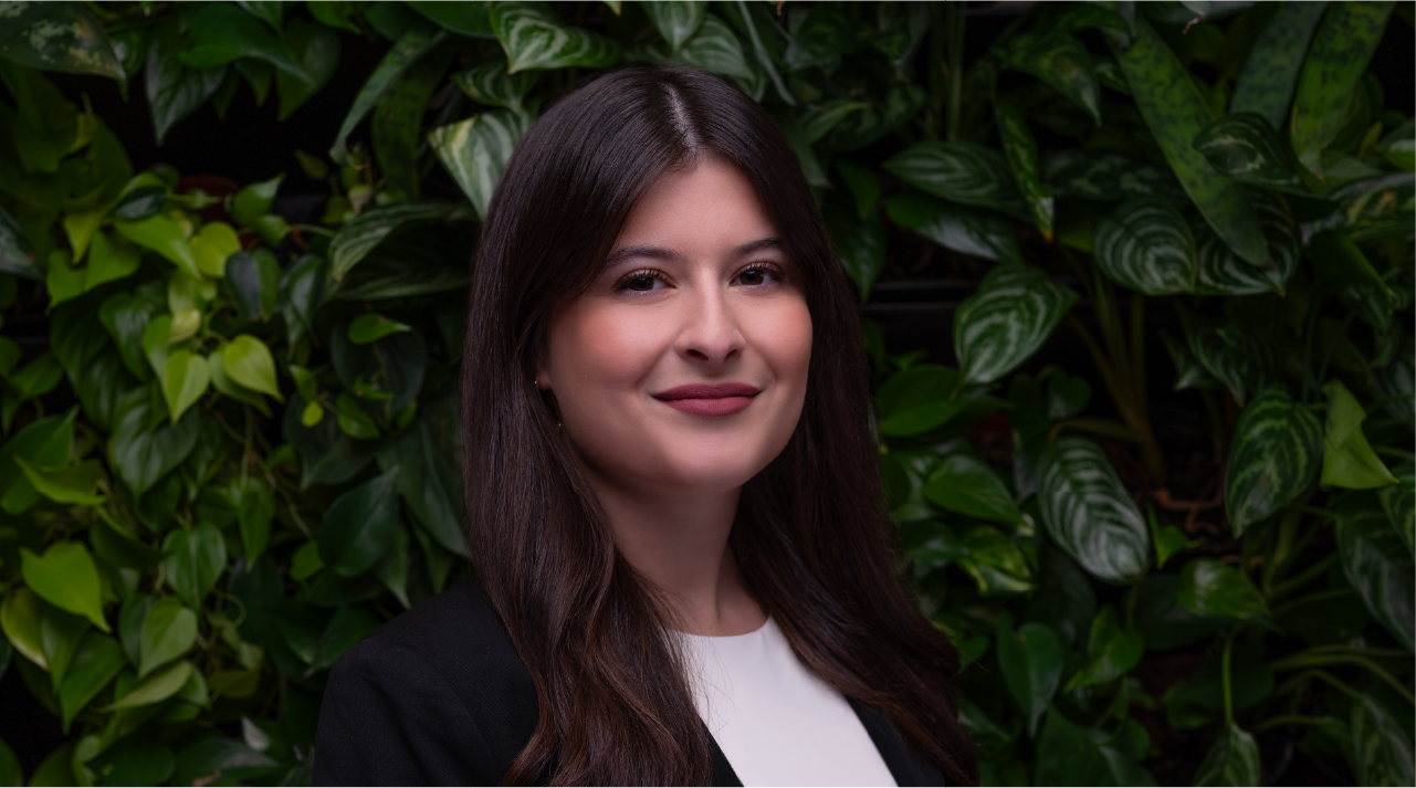Thalia Chaves wears a black jacket and smiles in front of a wall of green plants.