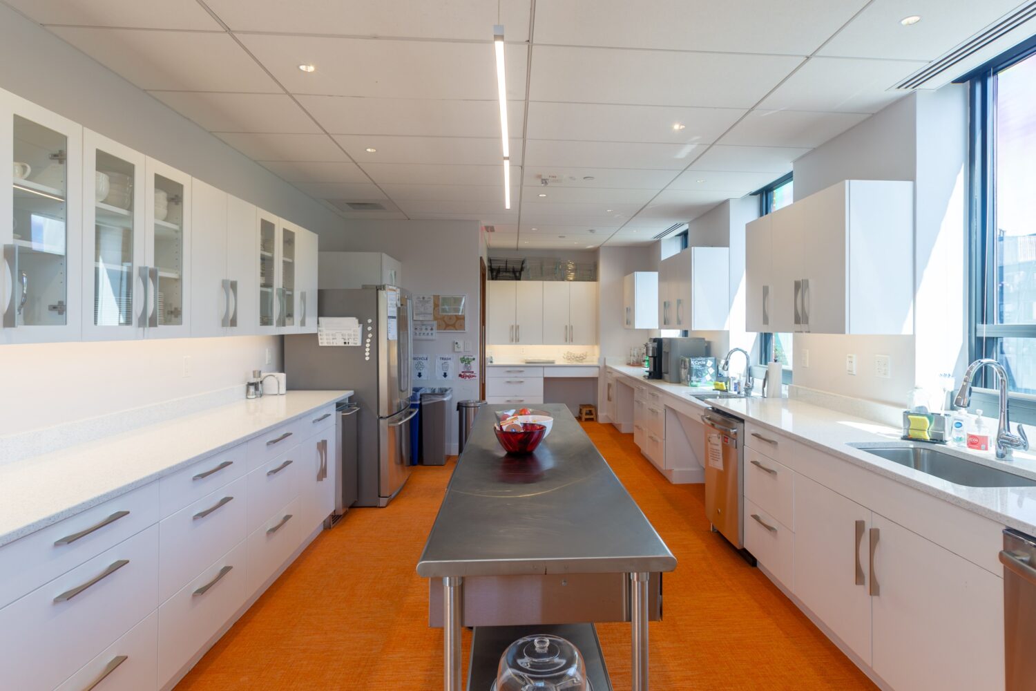Large kitchen with white cabinetry, orange flooring, and a steel table in the center.