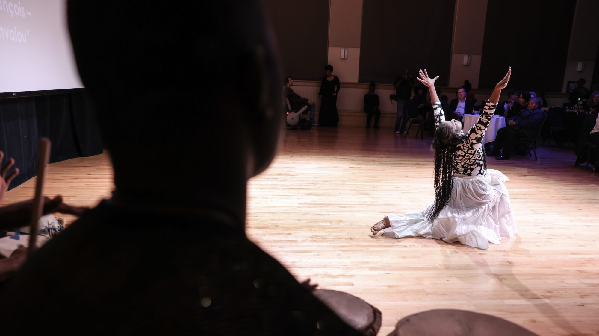A dancer in a white skirt poses in the foreground. A drummer watches on from the background.