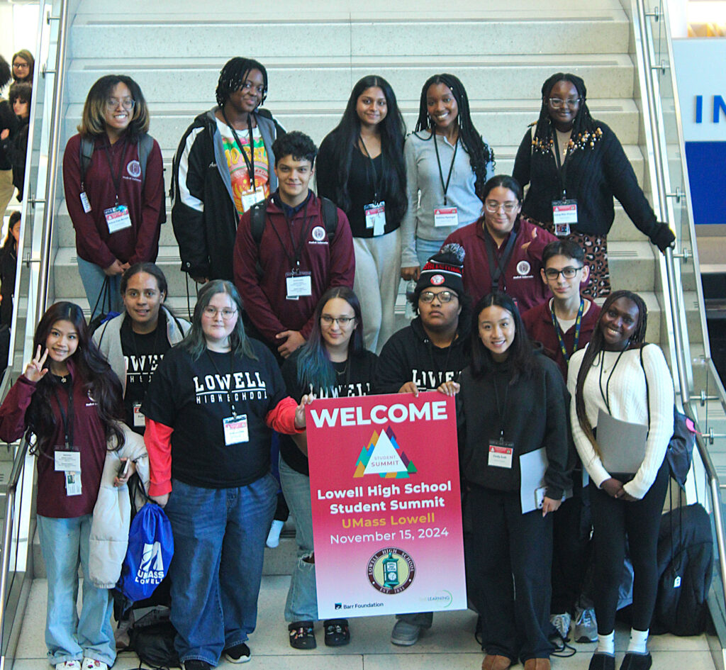 A group of high school students pose on a staircase, holding a sign that reads "welcome, Lowell High School Student Summit".