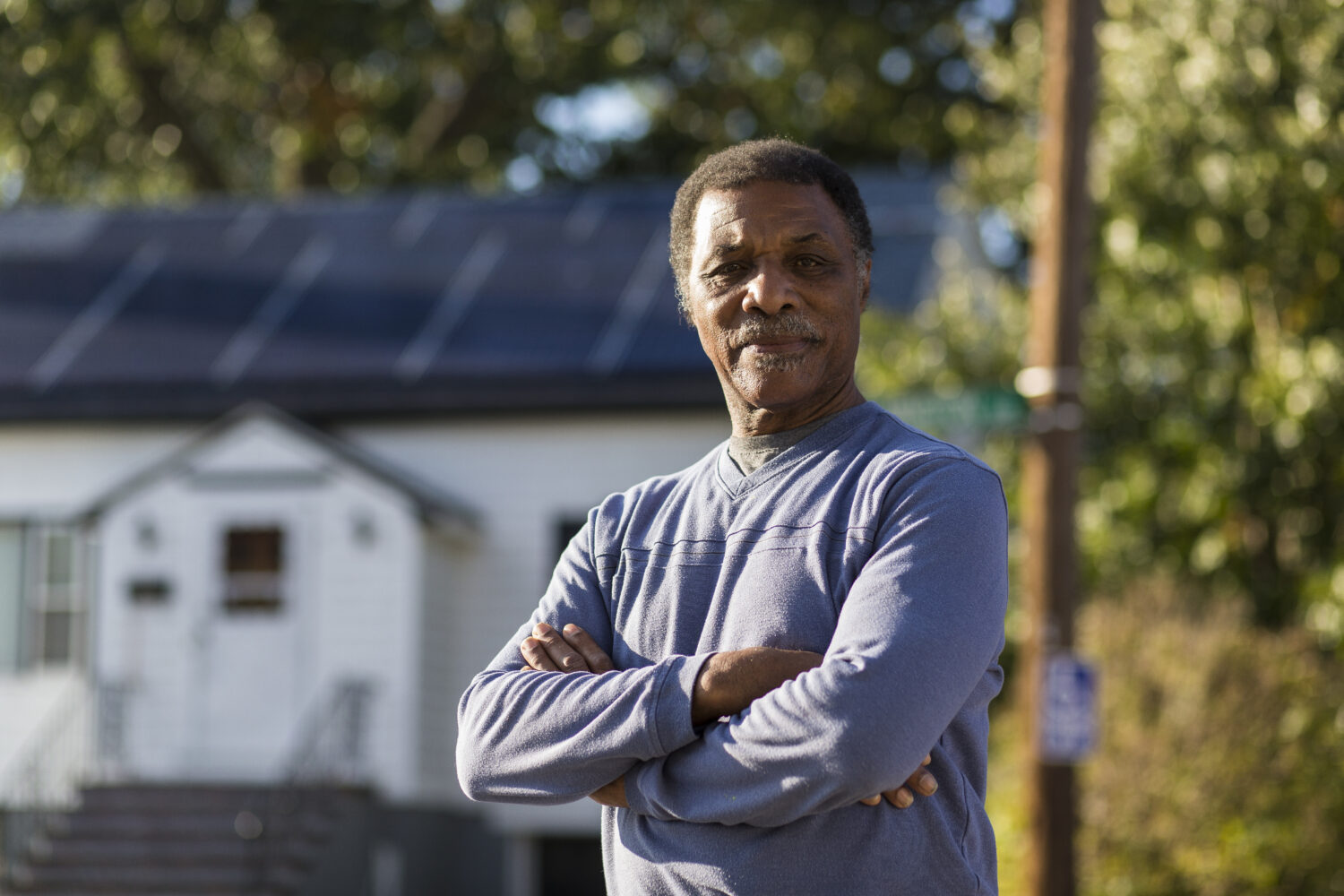 Older man stands in front of a house with solar panels on the room. He looks proud and has his arms crossed in front of his chest.