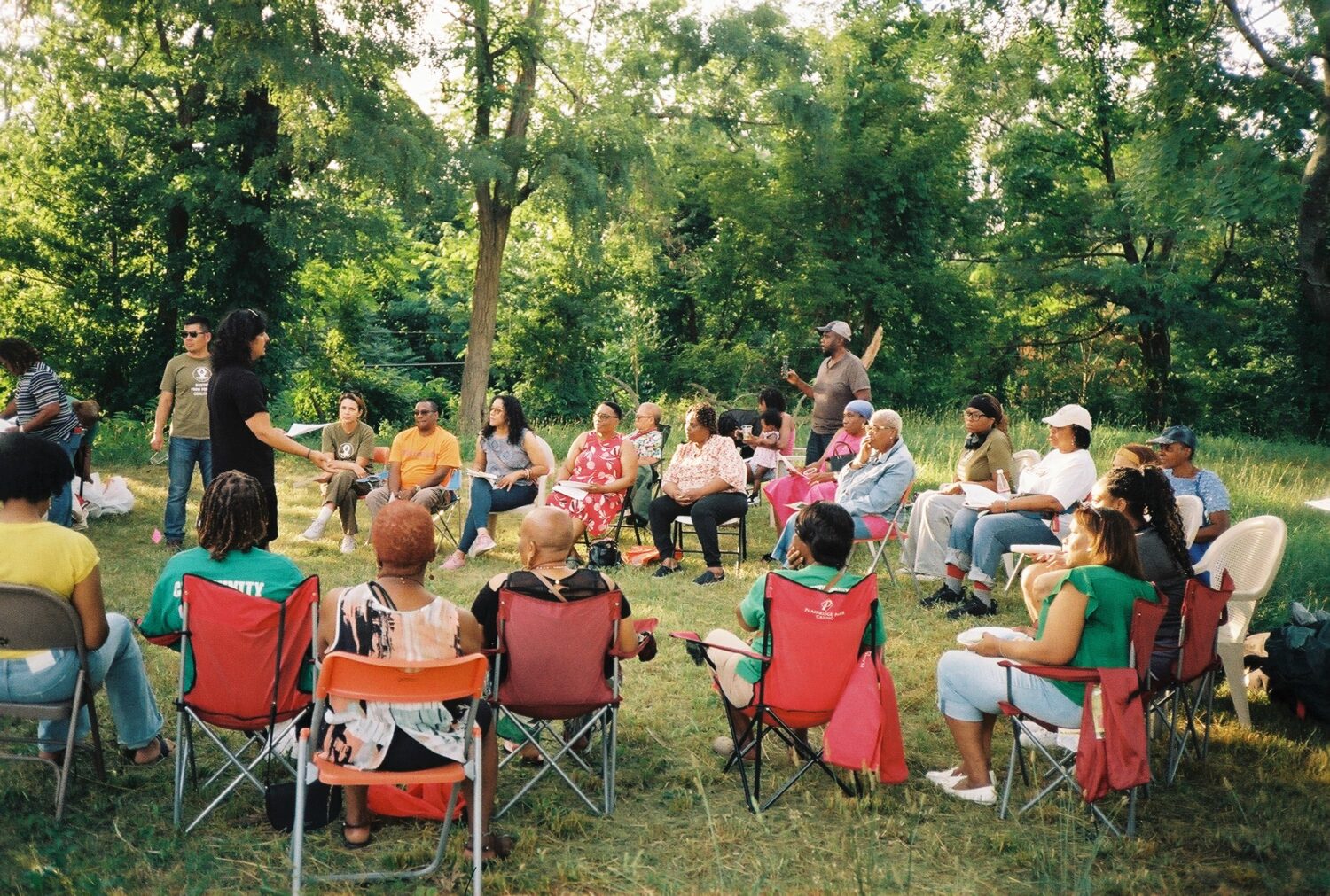 A group of twenty people sit in a circle in colorful camping chairs in calm setting with green grass and trees setting.
